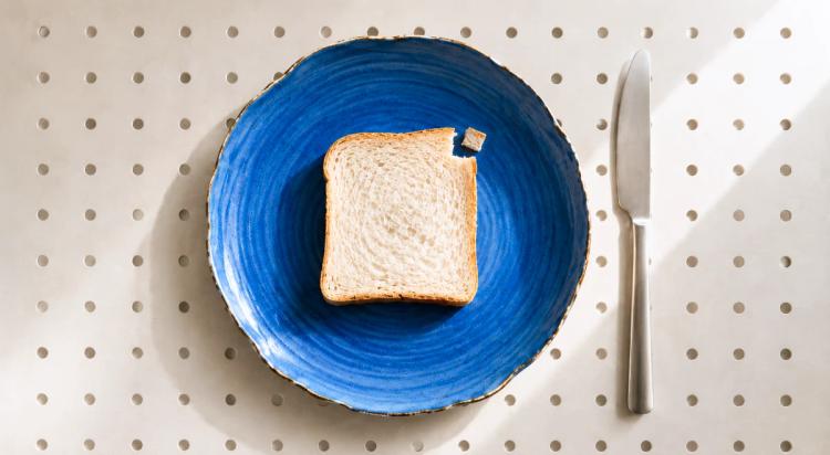 Rebanada de pan blanco sobre un plato azul con un cuchillo al lado, vista desde arriba sobre un fondo beige punteado, ilustrando una comida sencilla y concentrada como metáfora de microaprendizaje.